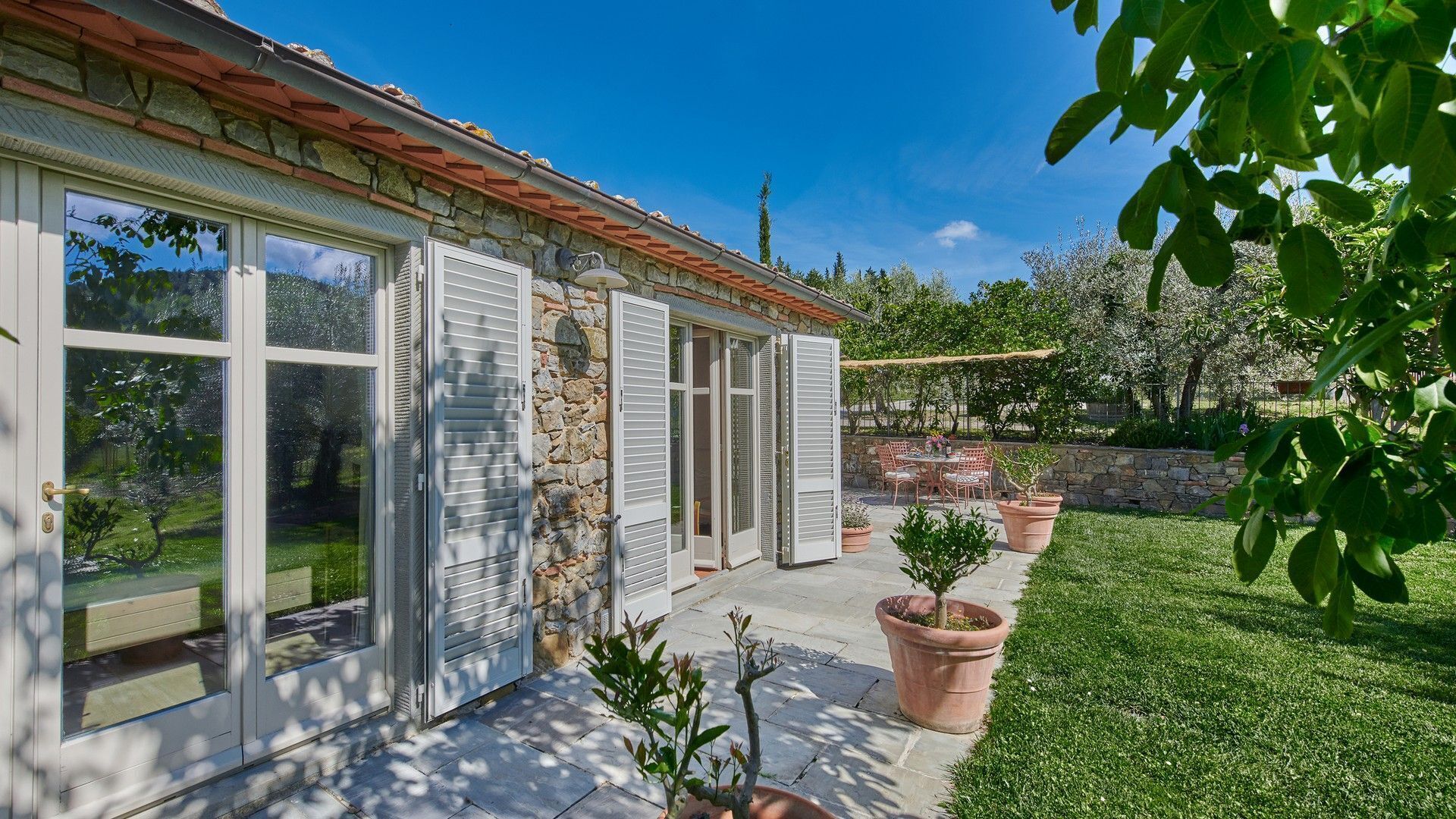 Una casa amb façana de pedra, grans finestres i un pati amb mobiliari d'exterior.
