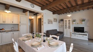 A dining area with a table set for four, a kitchen with white cabinets, and a living area with a TV and shelves. - Casa di Campagna Pianelli, Pergine Valdarno, Arezzo and Cortona (Pergine Valdarno)