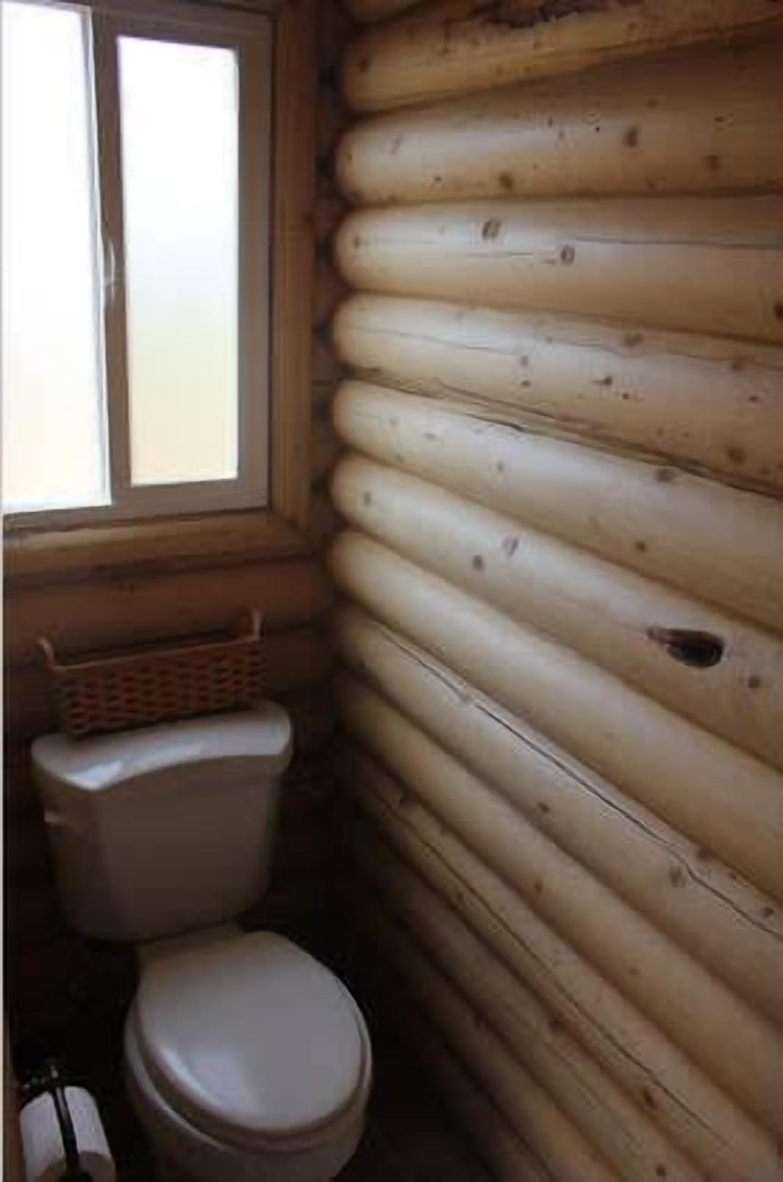 A log cabin bathroom with a toilet, a window, and a basket on the floor.