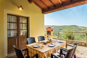 A dining area with a table set for two, a wooden door, and a view of a green hillside.