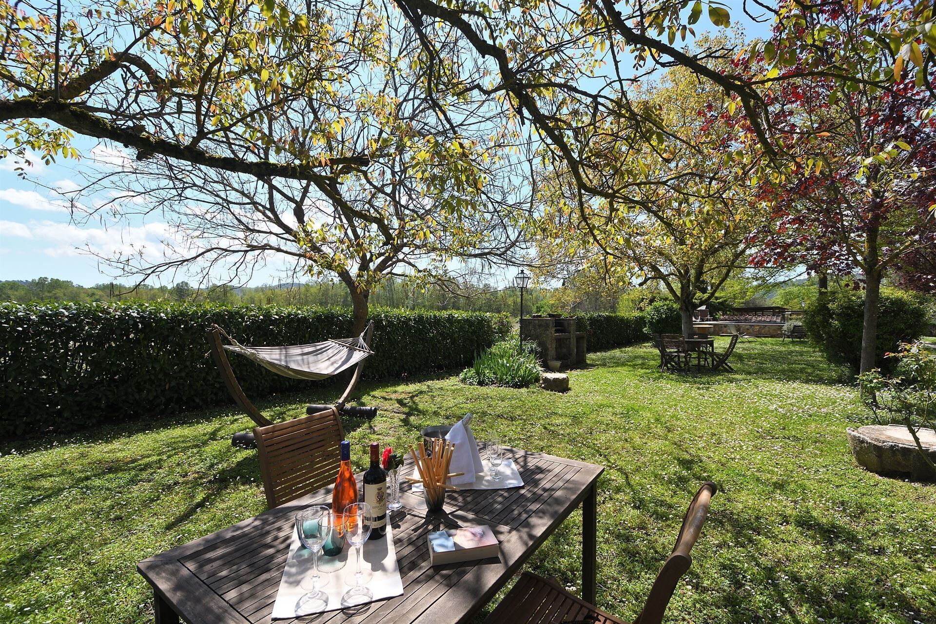 An outdoor dining area with a wooden table set for two, surrounded by lush greenery and trees.