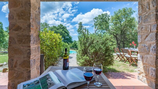 Uma pérgola de pedra com vista para uma piscina e árvores, uma garrafa de vinho, dois copos de vinho e um menu sobre a mesa.