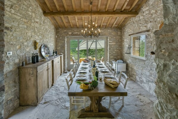 A rustic stone-walled dining area with a wooden table set for a meal, a chandelier, and a view of greenery outside.