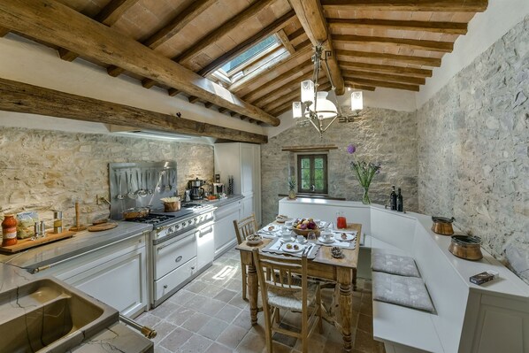 A rustic kitchen with stone walls, wooden beams, and a dining area with a bench.