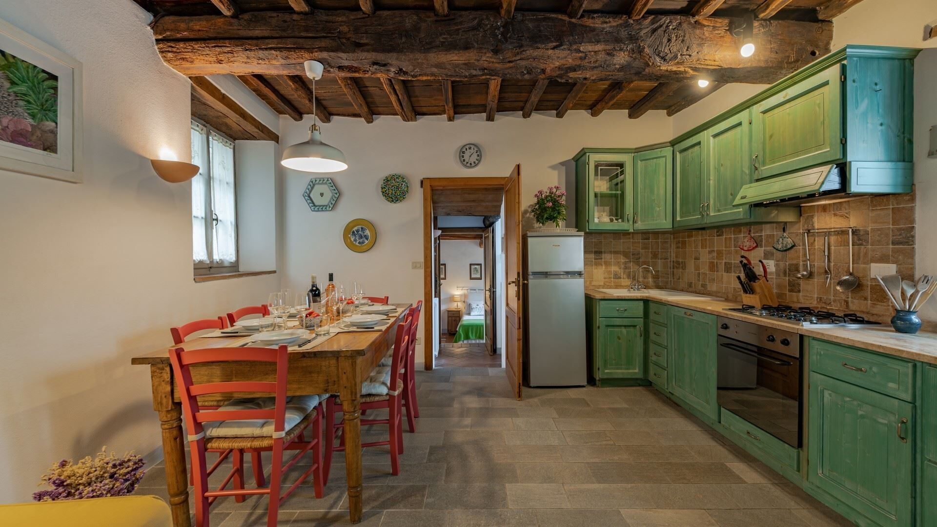 Eine rustikale Küche mit grünen Schränken, einem Holztisch mit roten Stühlen und einem sichtbaren Kühlschrank.