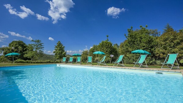 A clear blue swimming pool with lounge chairs and umbrellas, surrounded by trees and a clear sky.