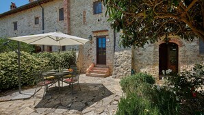 A stone building with a patio area featuring a table and chairs under an umbrella, surrounded by greenery and a tree. - Pinturicchio, Pretaccione, Castelnuovo Berardenga, Siena and Chianti (Castelnuovo Berardenga)