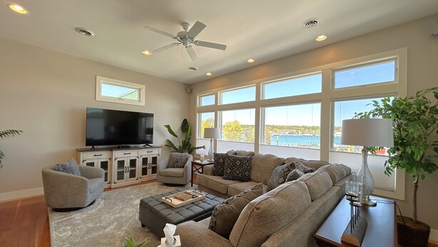 A spacious living room with a large flat-screen TV, comfortable seating, a ceiling fan, and large windows offering a view of the outdoors. - St Ignace Loft - Downtown St Ignace (Saint Ignace)