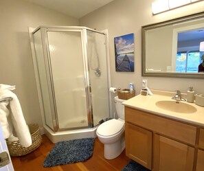 A bathroom with a glass shower enclosure, a toilet, a sink with a mirror, and a blue rug on the floor. - St Ignace Loft - Downtown St Ignace (Saint Ignace)