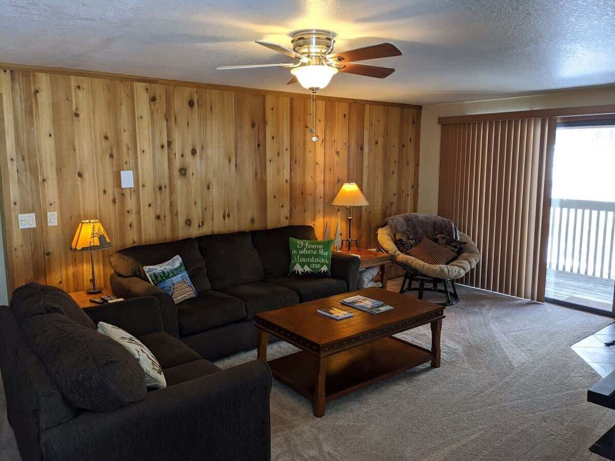 A cozy living room with a wooden wall, a ceiling fan, a sofa, a chair, a coffee table, and a lamp.