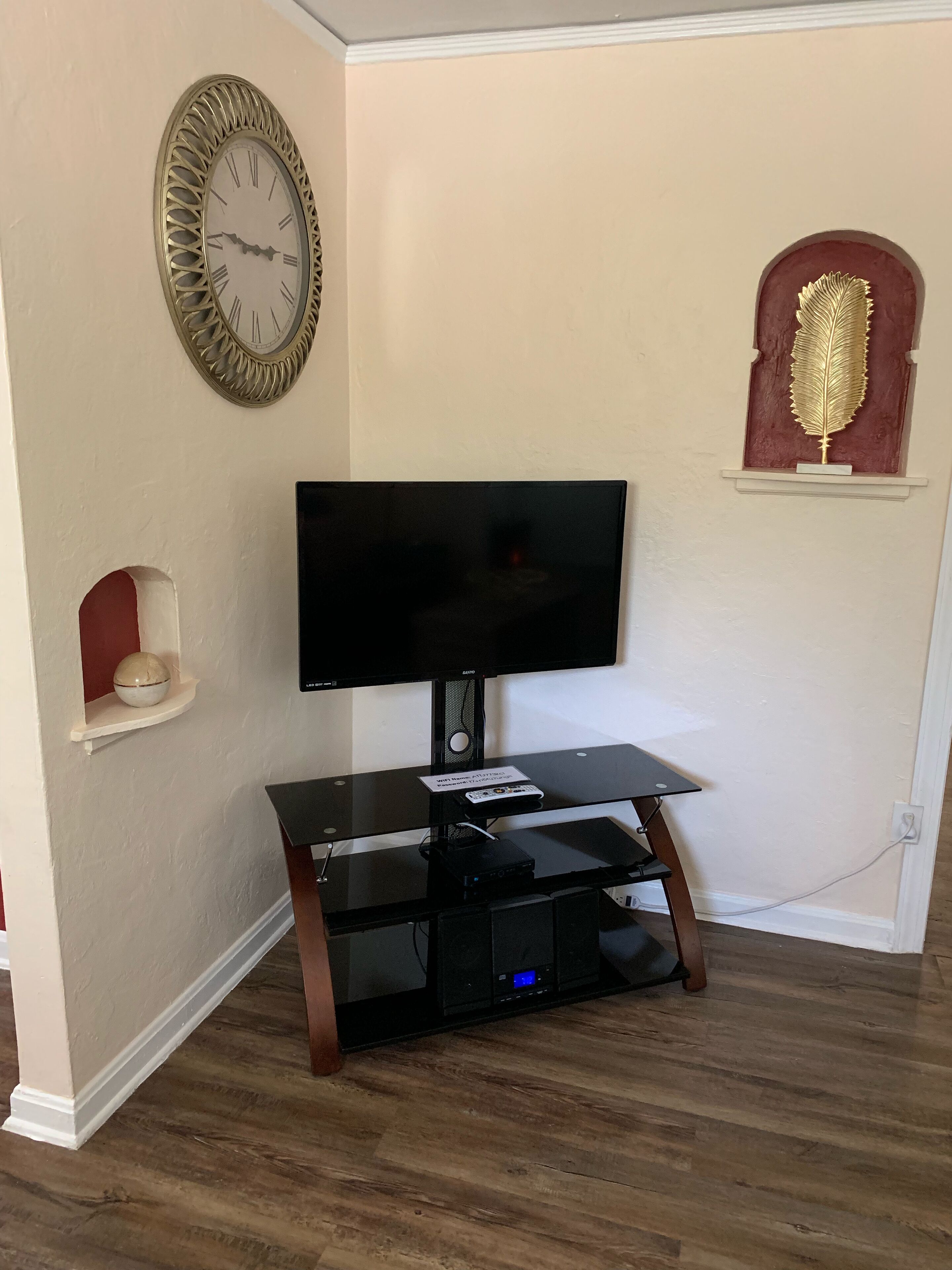 A living room with a flat-screen TV on a black stand, a decorative clock, and a small shelf with a decorative item.