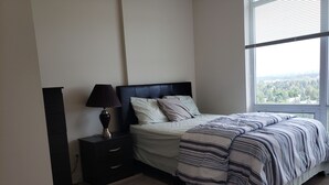 A bedroom with a bed, bedside table, lamp, and a view through a window. - A perfect escape from regular hotels? Check out this attractive Condo unit. (Calgary)