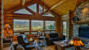 L’intérieur d’une cabane en rondins est agrémenté d’une cheminée, de grandes fenêtres offrant une vue sur les montagnes, de meubles en cuir et d’un lustre.