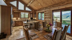 Une cuisine spacieuse aménagée dans une cabane en rondins, avec un grand îlot central, des armoires en bois et un coin repas comprenant six chaises et une table dressée pour six personnes.