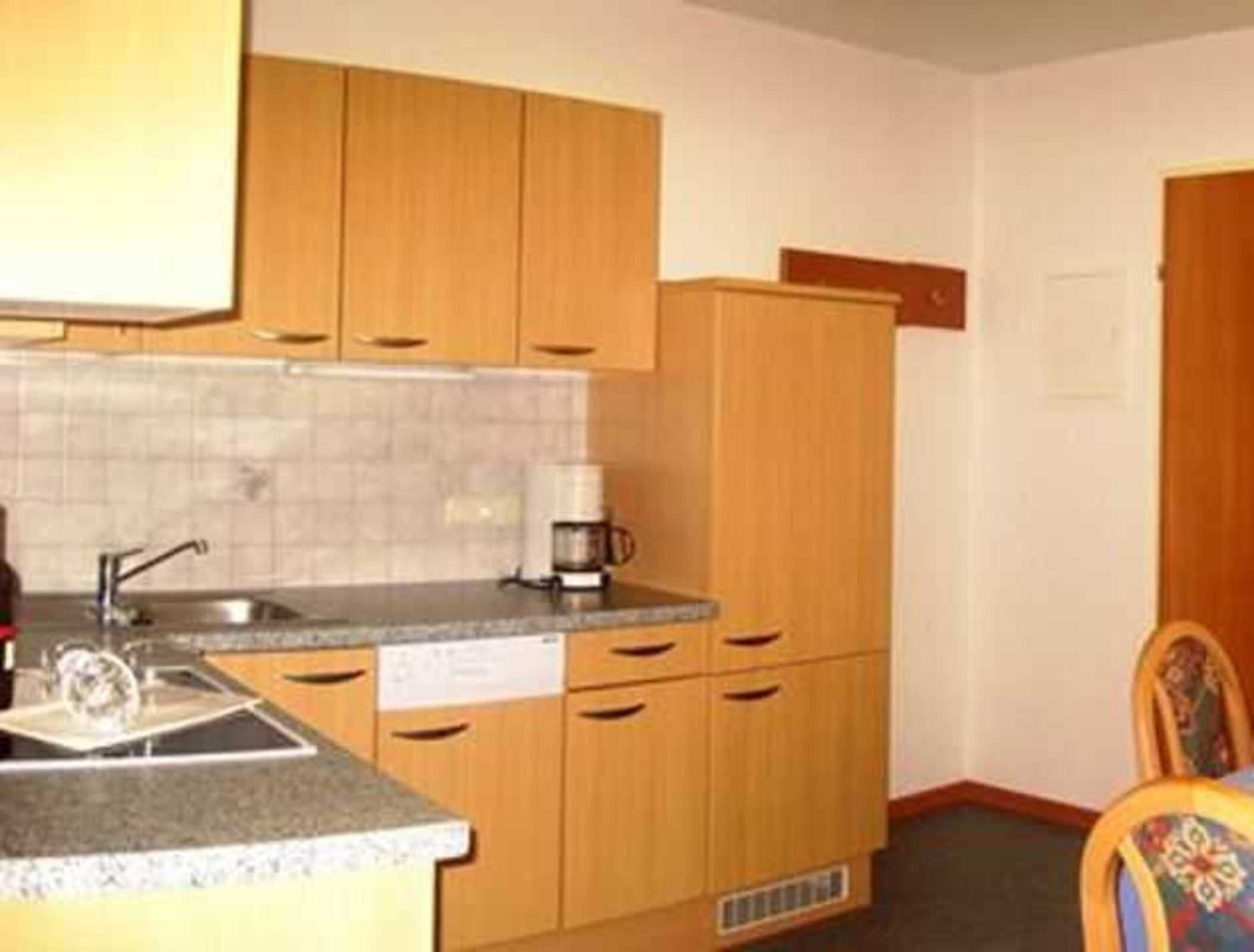A compact kitchen with wooden cabinets, a sink, a coffee maker, and a dining area with a chair.