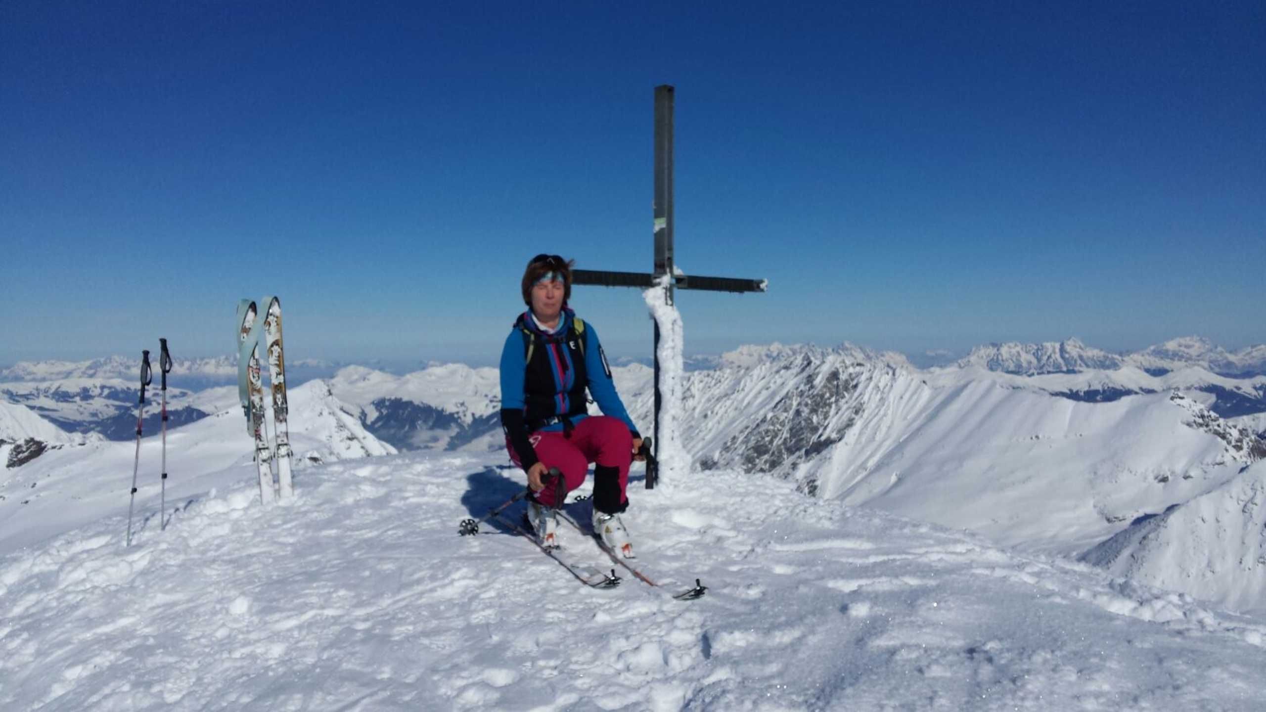 A person sitting on a snowy mountain with skis and poles, a cross, and a clear blue sky in the background.