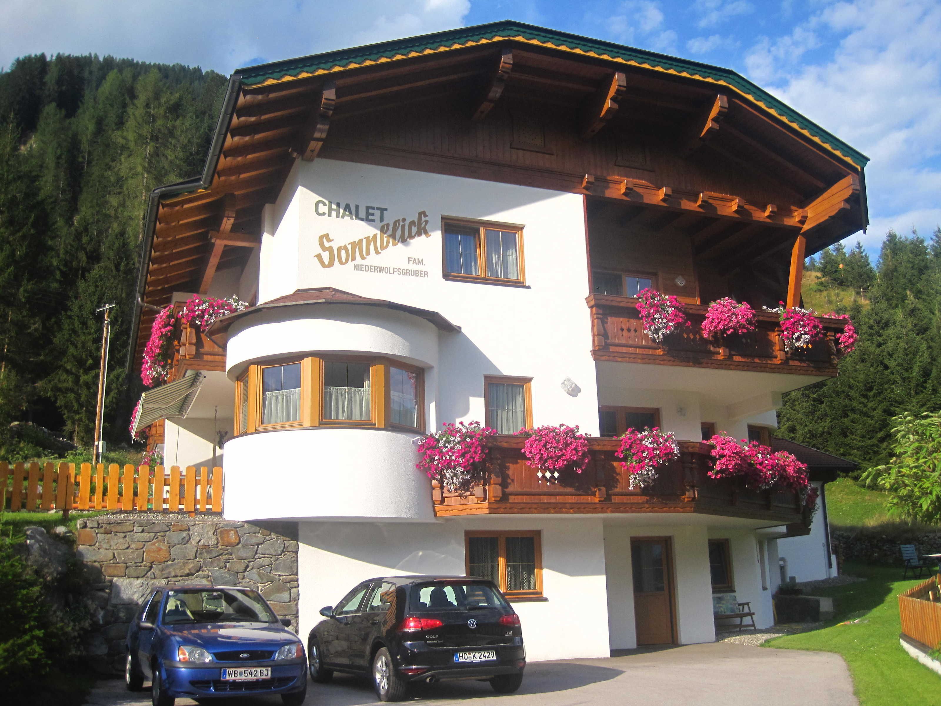 Una cabaña con un cartel que dice 'CHALET Sonnablick FAM. Niederwolfsgruber', con balcones de madera con jardineras, un muro de piedra y coches aparcados en la parte delantera.