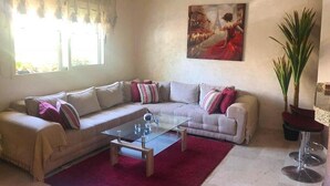 Un salon avec un canapé beige, une table basse en verre et un tapis rouge. Il y a un tableau au mur et une plante en pot.