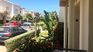Un quartier résidentiel avec des voitures garées, un jardin bien entretenu avec des palmiers et des fleurs rouges, et un bâtiment blanc orné de colonnes.