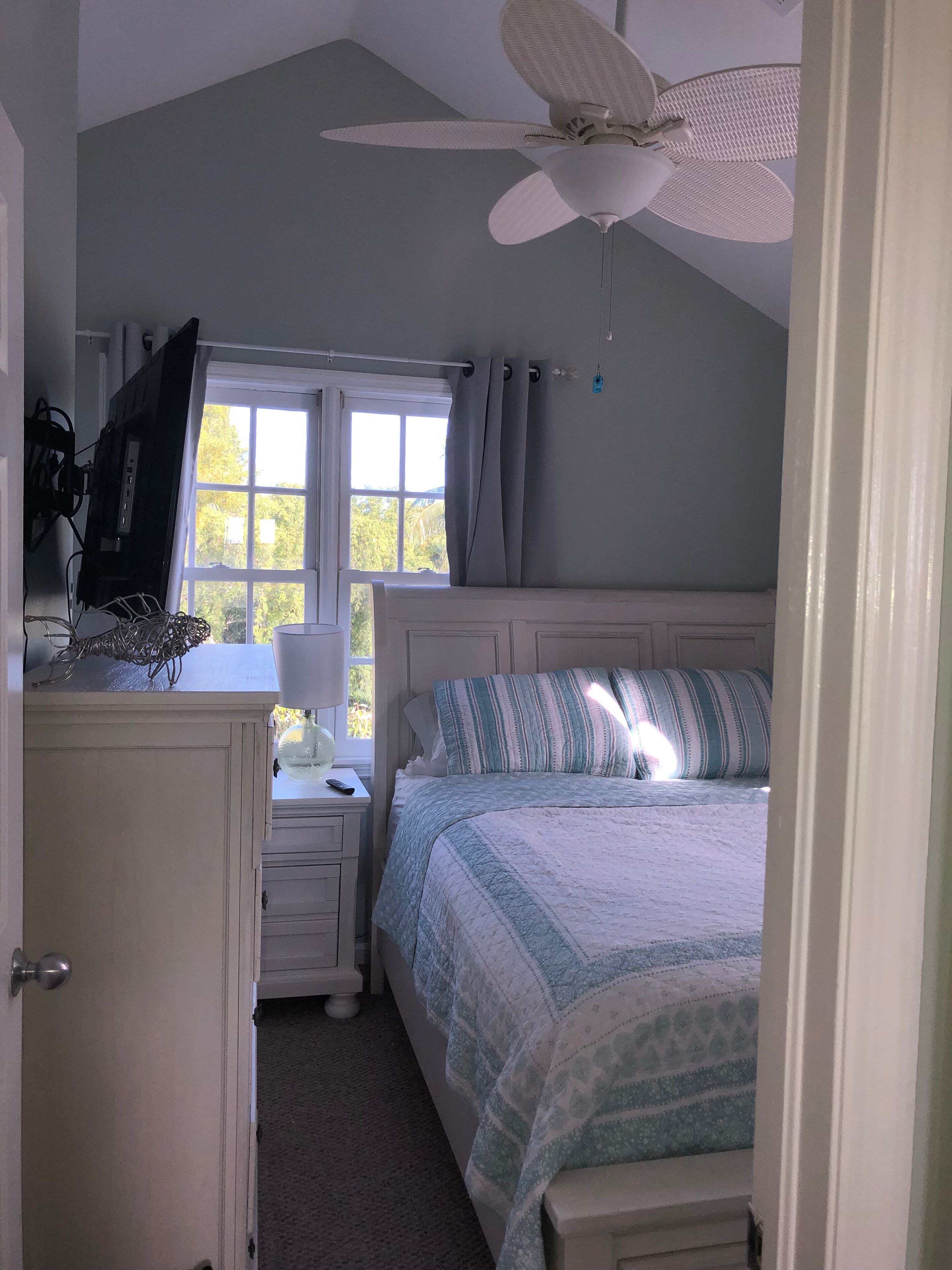 A bedroom with a bed, a ceiling fan, a window with curtains, a desk with a lamp, and a TV on a stand.