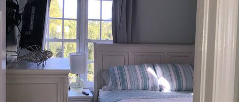 A bedroom with a bed, a ceiling fan, a window with curtains, a desk with a lamp, and a TV on a stand.