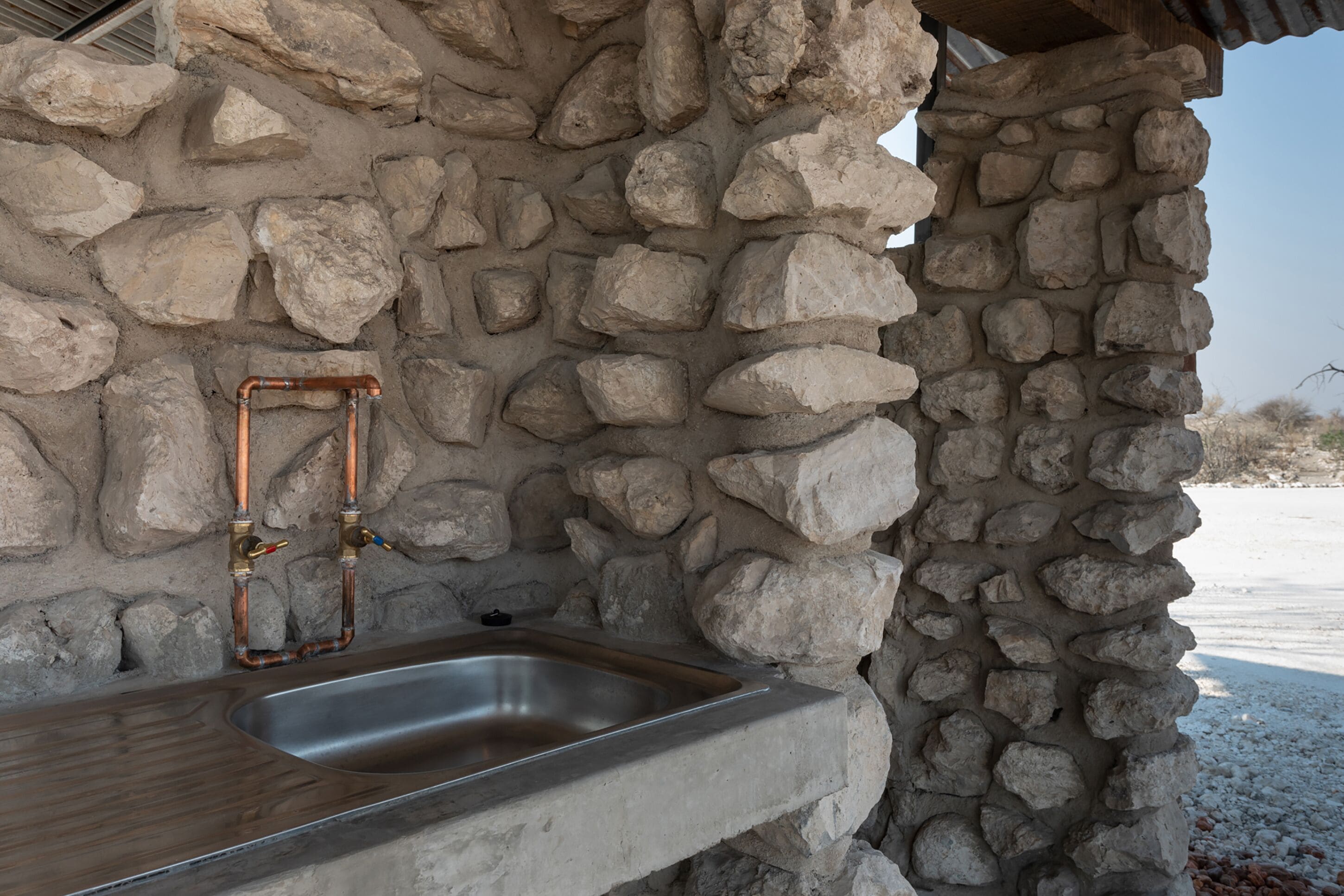 Pared exterior de piedra con un lavamanos de metal y tuberías de cobre.