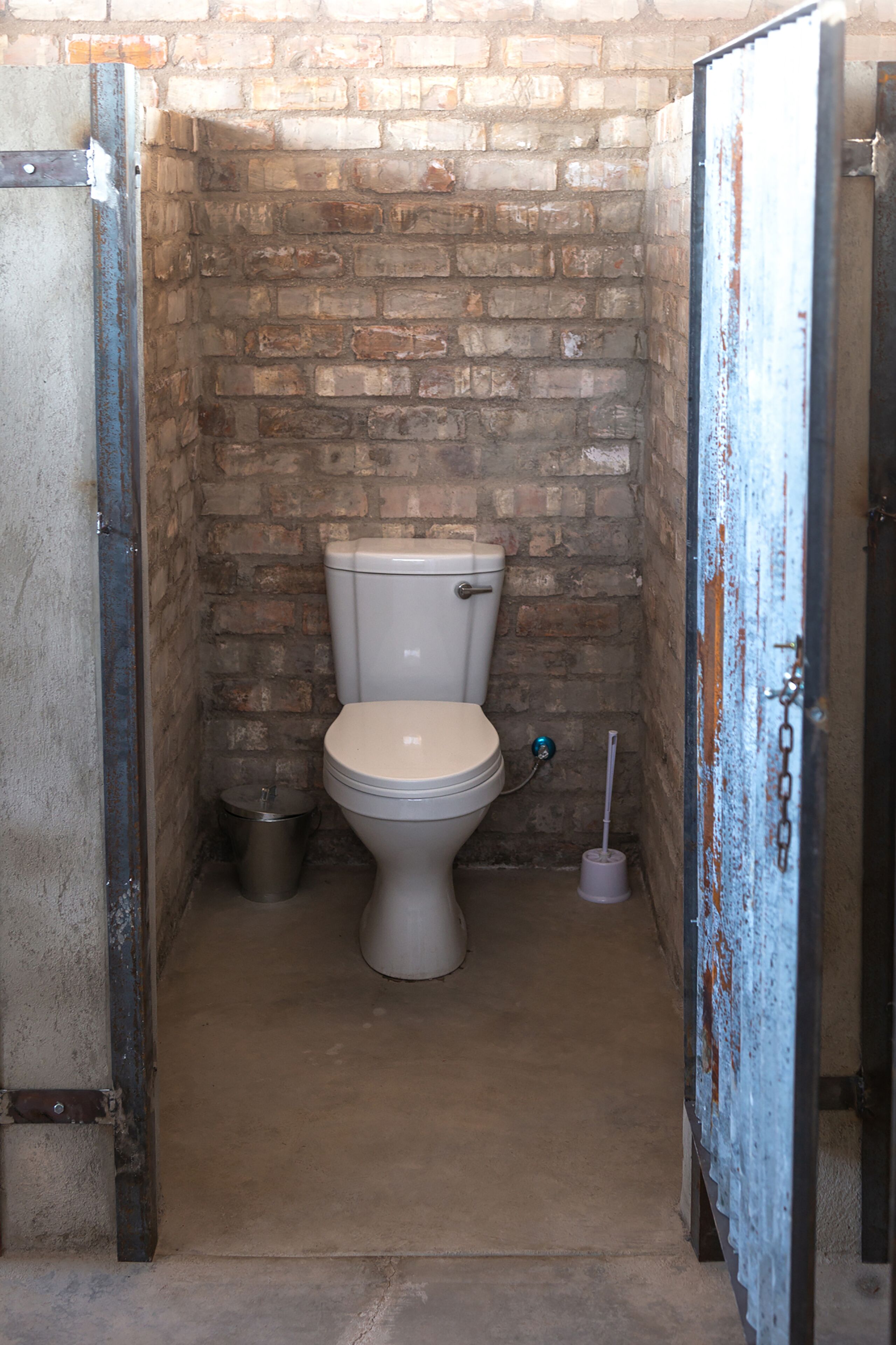 Een kleine, afgesloten badkamer met een toilet, een prullenbak en een toiletborstel.