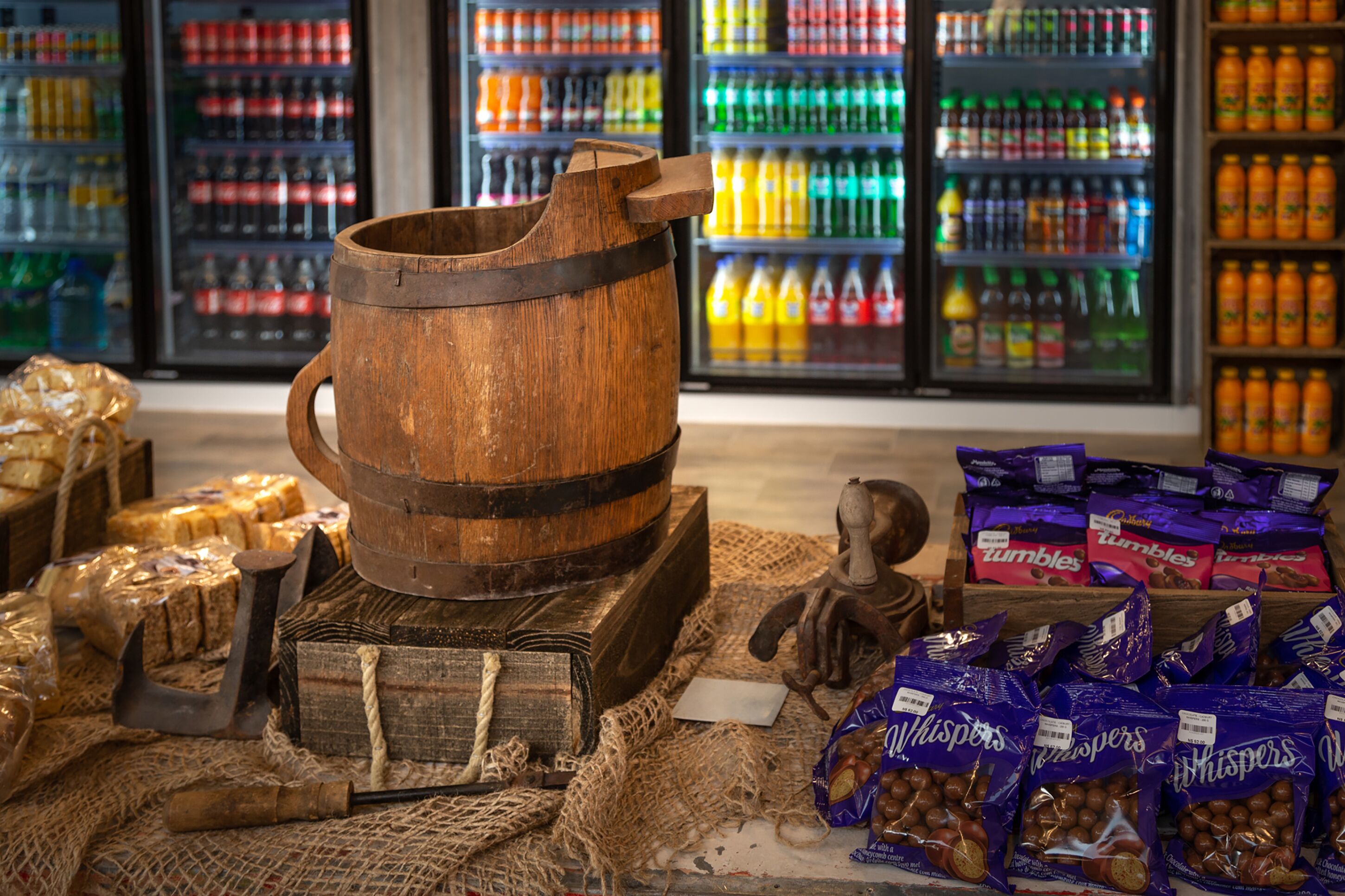 A rustic wooden barrel, burlap sacks, and chocolate packages in a store with colorful beverage shelves in the background.