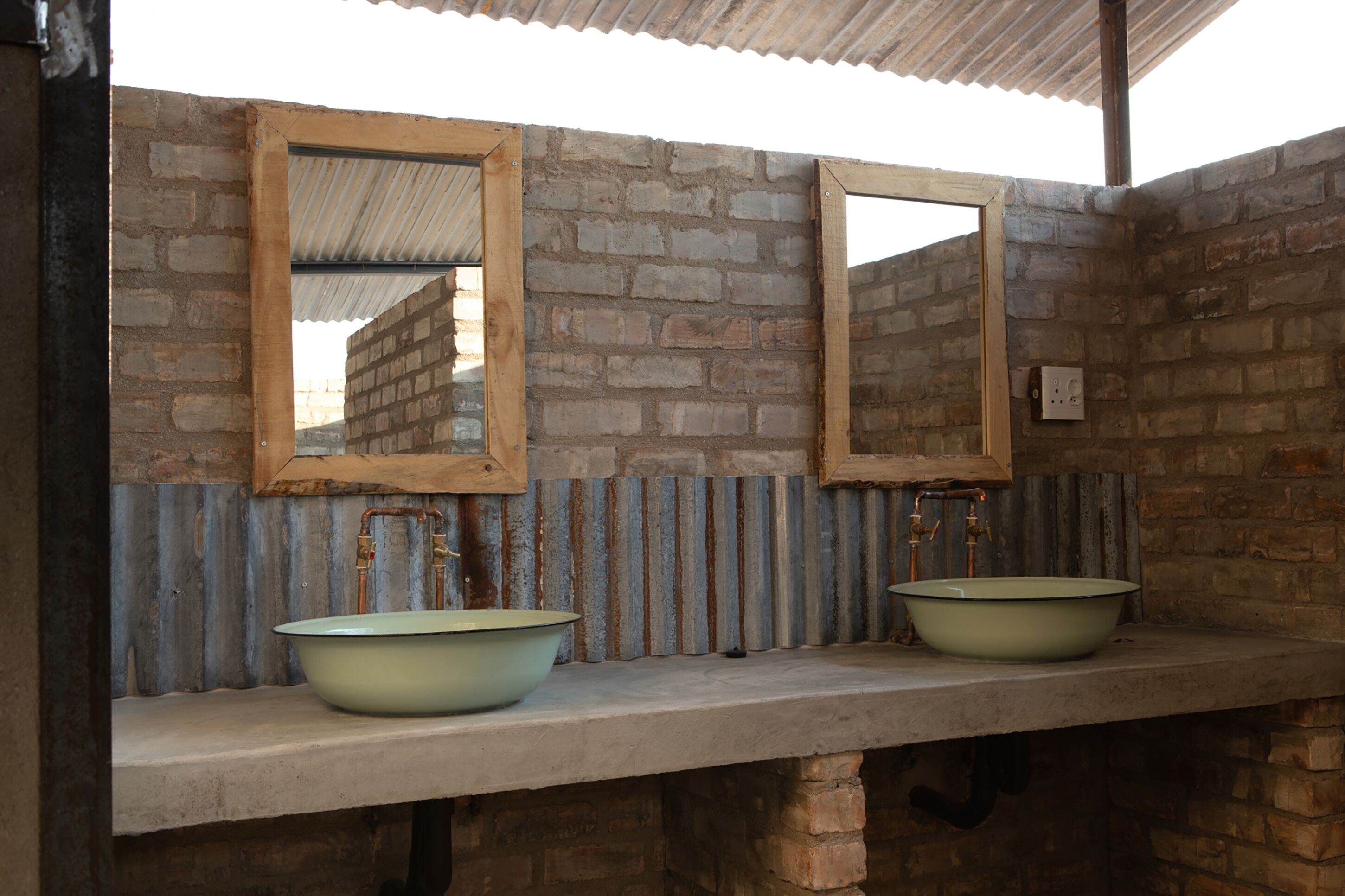A bathroom with two sinks, each with a mirror above. The walls are made of bricks, and there is a corrugated metal roof.