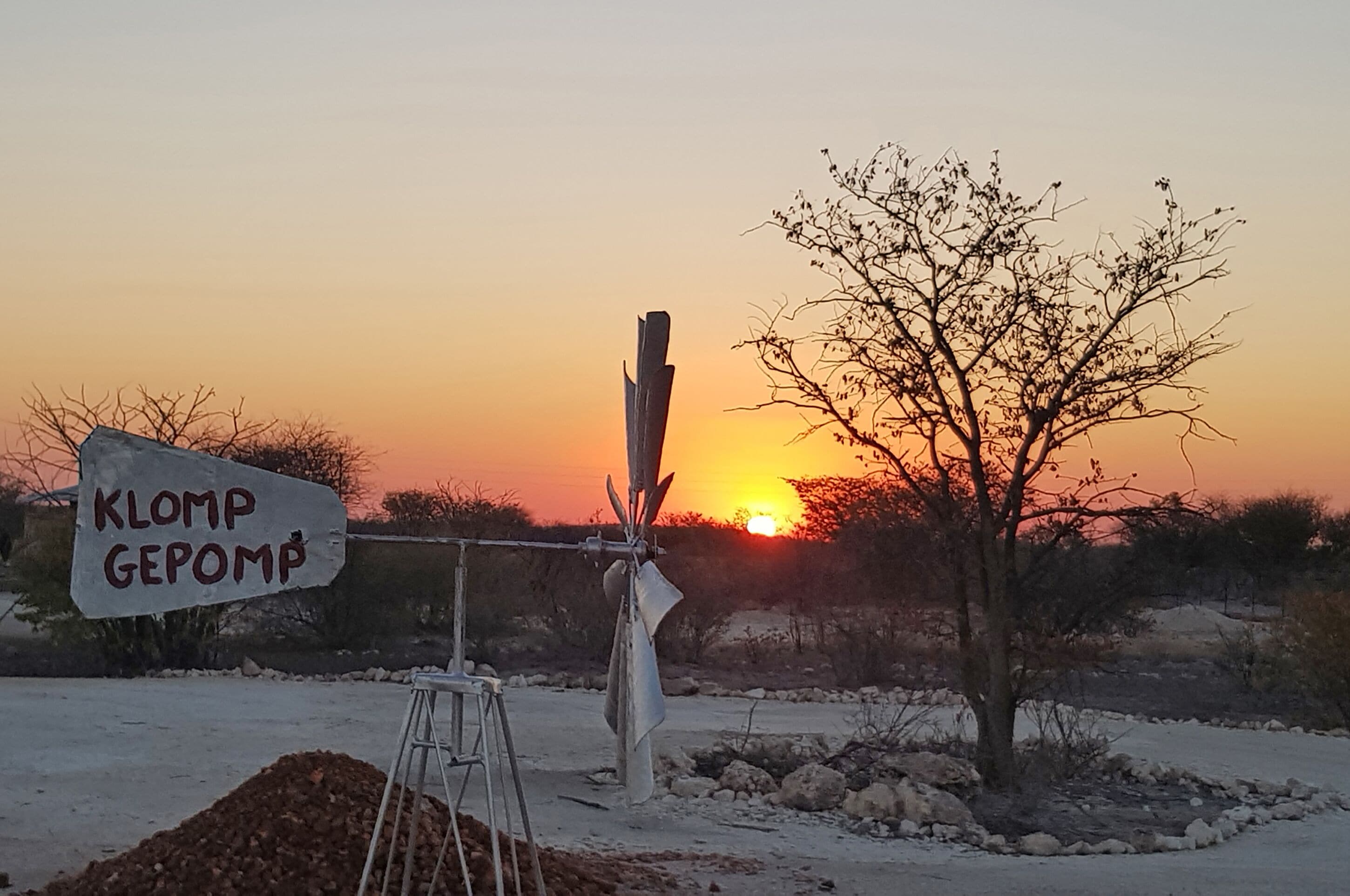 A sign with text, a metal structure, a tree, and a sunset in the background.
