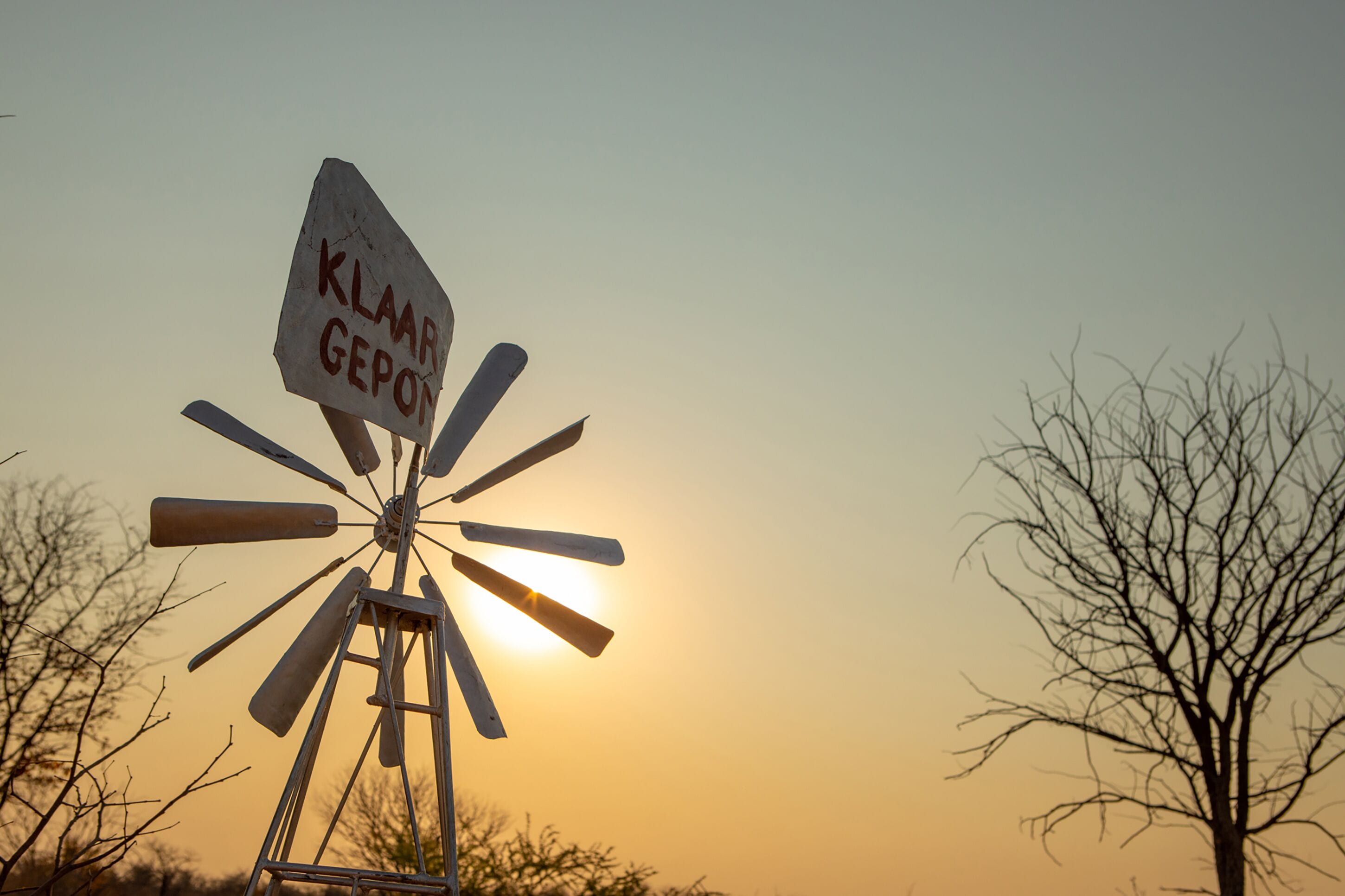 A windmill with a sign that reads "KLAAR GEOPOT" against a sunset sky.