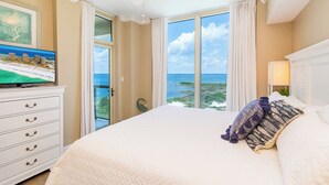 Habitación de hotel con una cama grande, un televisor en una cómoda y vista al océano a través de amplias ventanas.