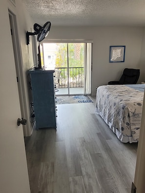 Une chambre à coucher avec un lit, un bureau équipé d’un ventilateur, une chaise et une vue sur l’extérieur grâce à une porte coulissante.