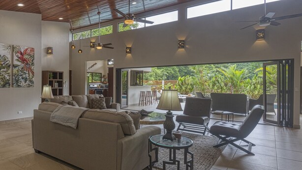 A spacious living area with a sofa, chairs, and a coffee table. There are ceiling fans and wall-mounted lights. The room has large windows offering a view of greenery outside. - New Luxury Home, private pool! 300 steps to beach, 8 min. walk to bars & dining (Dominical de OSA)
