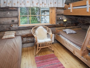 A log cabin room with a wooden bunk bed, a wicker chair with a fur throw, a window with a view of trees, and a patterned rug.