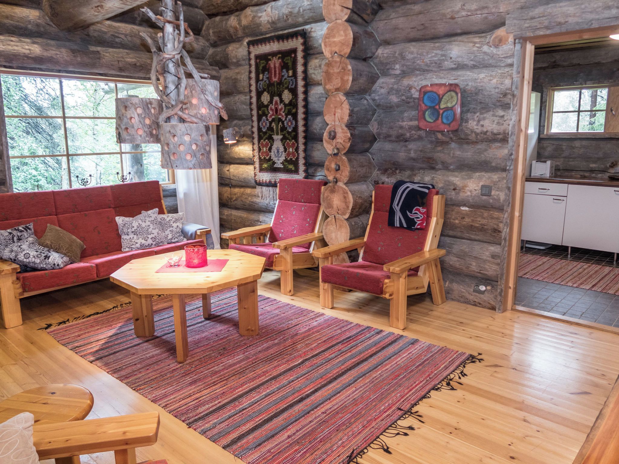 A log cabin interior with a wooden dining table, red upholstered chairs, a patterned rug, and a window with a view of trees.