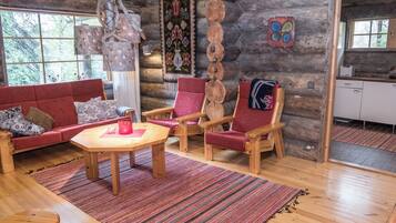 A log cabin interior with a wooden dining table, red upholstered chairs, a patterned rug, and a window with a view of trees.