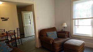 Un salon avec un canapé marron, un pouf et des coussins à motifs. On y trouve une table à manger avec des chaises et une lampe sur une table d’appoint.
