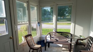 Une véranda donnant sur une rue et sur un espace verdoyant, avec une chaise en bois, une chaise en plastique et une petite table.