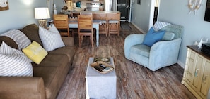 A living room with a brown sofa, a blue armchair, a dining table with chairs, and a kitchen in the background.