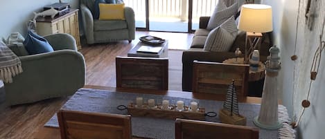 En stue med spisebord, stole, sofaer og en balkon med et skilt, der står 'BEACH HOUSE'.