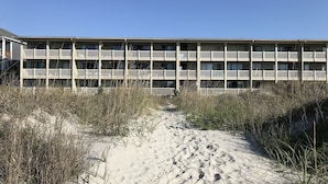 Un edificio de varios pisos con balcones, un camino de arena que conduce hacia él y vegetación circundante.