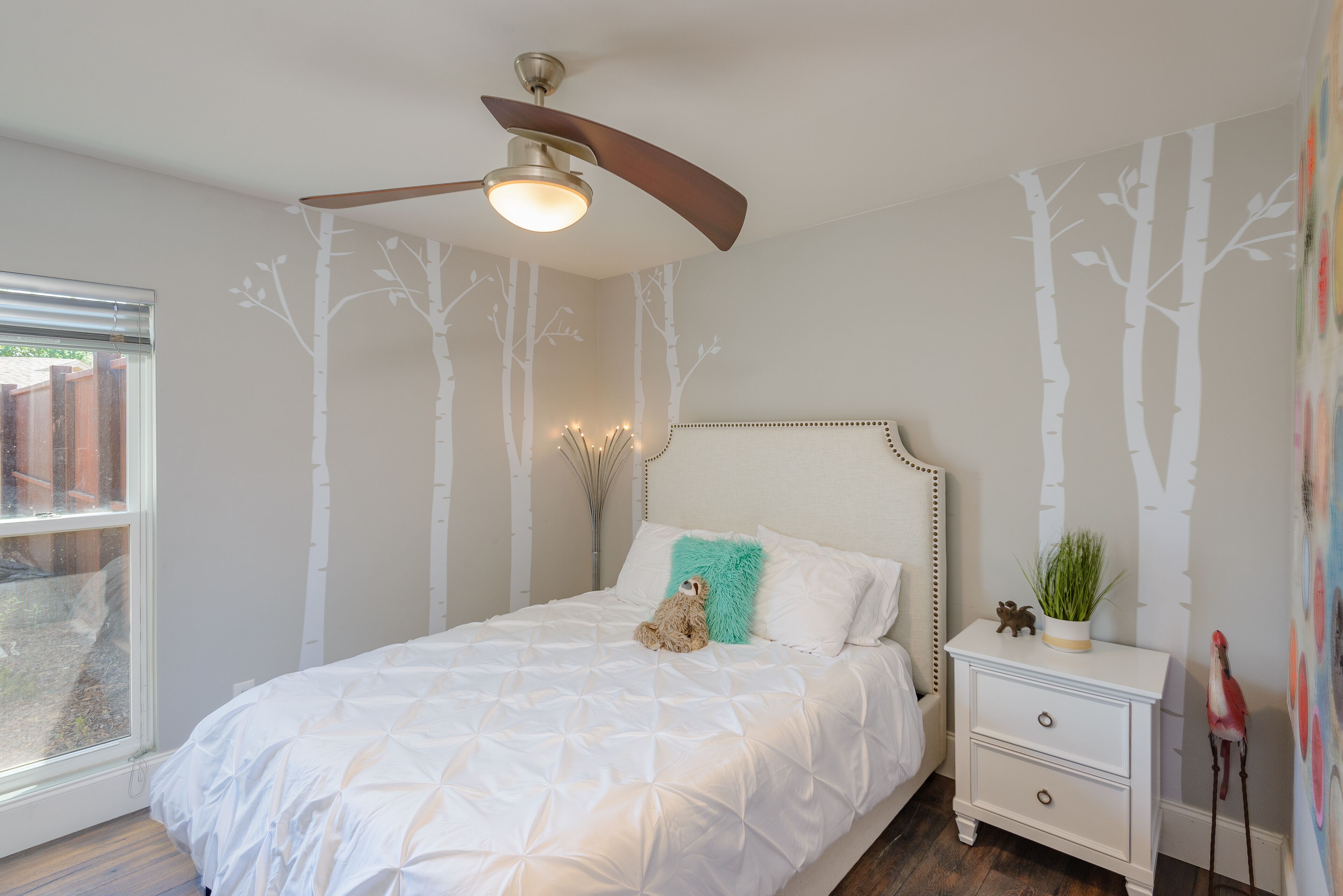 A bedroom with a bed, a nightstand, a ceiling fan, and wall decals of trees.