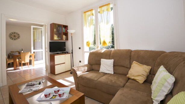 A living room with a brown sofa, a coffee table, and a television. There is a dining area with a table and chairs visible through an open door. - Porto Letizia Mimosa 2, Porlezza, Italy (Porlezza)