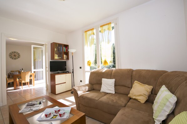 A living room with a brown sofa, a coffee table, and a television. There is a dining area with a table and chairs visible through an open door.