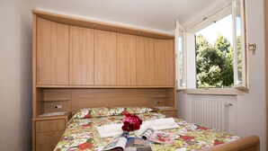 A bedroom with a bed, a wooden wardrobe, a nightstand, and a window with a view of trees. - Porto Letizia Mimosa 2, Porlezza, Italy (Porlezza)