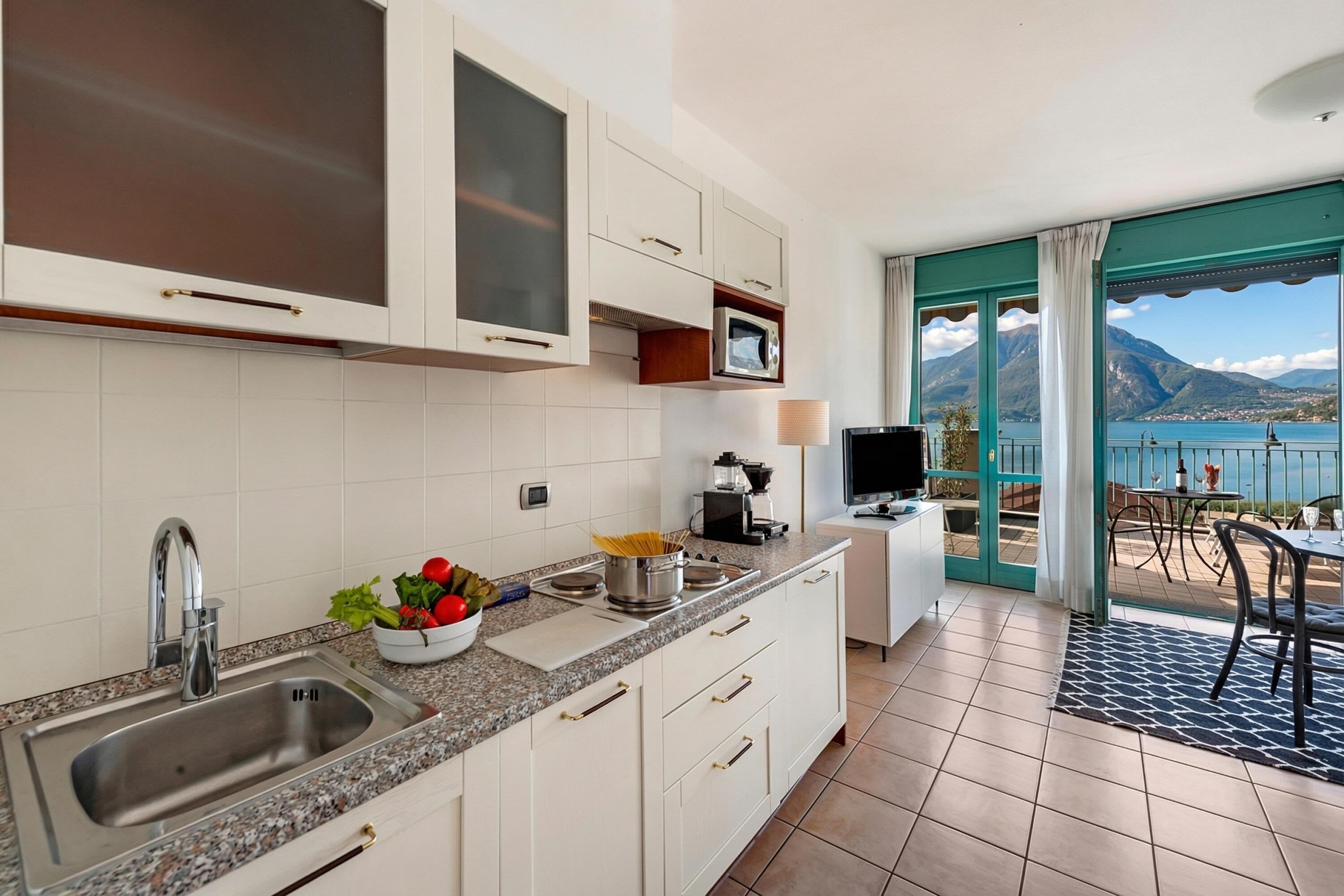 A modern kitchen with a sink, stove, and microwave, and a balcony with a view of the lake and mountains.