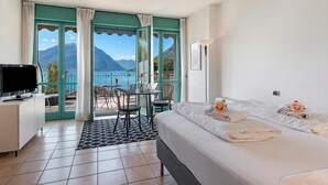 A hotel room with a bed, a TV, a desk, a chair, a table, and a view of the sea and mountains. - Porto Letizia P10, Porlezza, Italy (Porlezza)