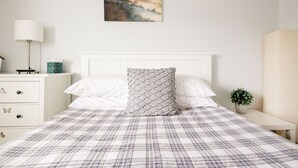 A bedroom with a plaid bedspread, white headboard, a bedside table with a lamp, a small plant, and a wall-mounted picture.