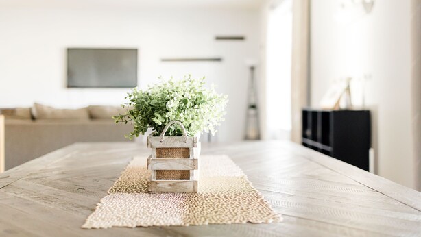 A wooden table with a woven placemat, a wicker basket with a plant, and a modern living room in the background.